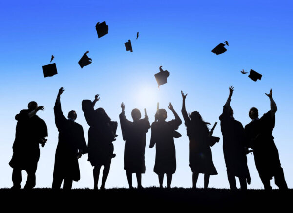 Silhouettes of graduates in caps and gowns tossing mortarboards into the air against a blue sky at sunset/bright sun behind them.