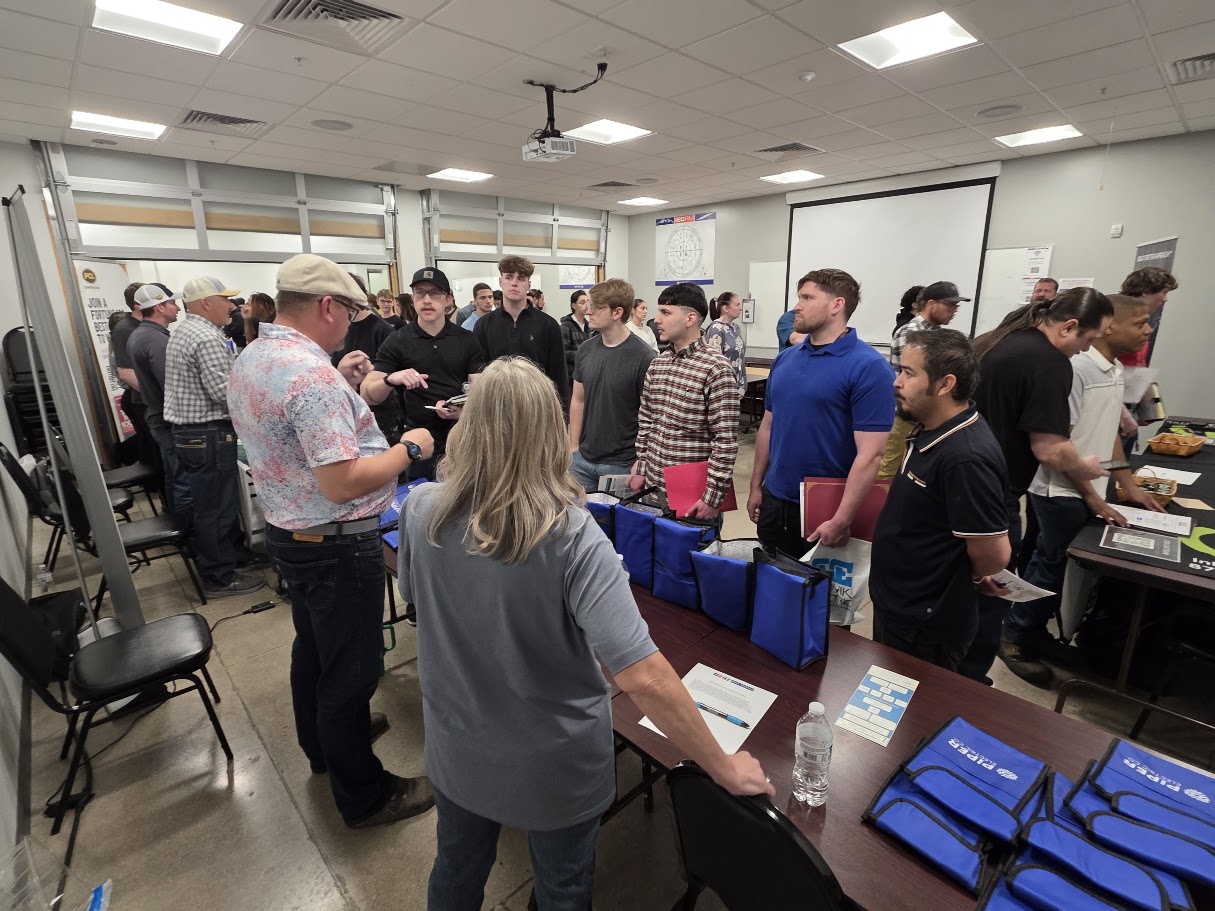 Group of job seekers lined up at a career fair, talking with recruiters at tables with blue tote bags on display.