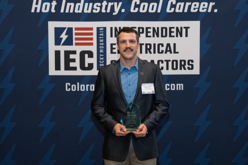 Man in a blazer holding a glass award, posing in front of an Independent Electrical Contractors banner at a conference.