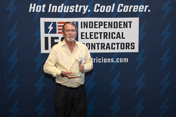 Man in a pale yellow shirt holding a glass award in front of a blue Independent Electrical Contractors banner.