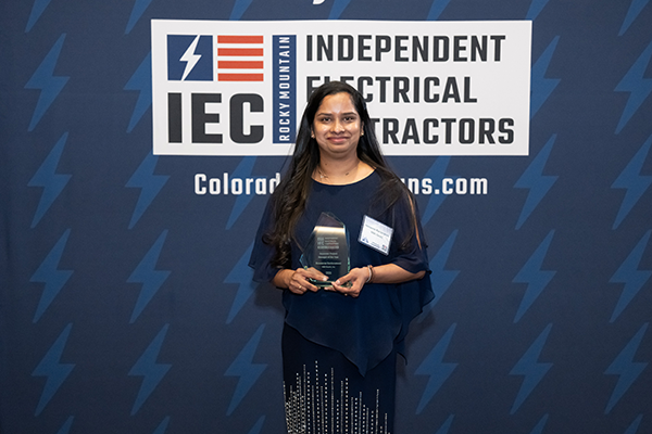 Woman in a dark blue dress holding a glass award in front of an IEC Independent Electrical Contractors backdrop with a lightning bolt pattern behind her.