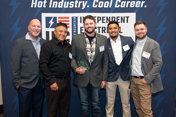 Five men in business attire pose together at an industry awards event; the man in the center holds a glass trophy, with a blue backdrop and event signage behind them.