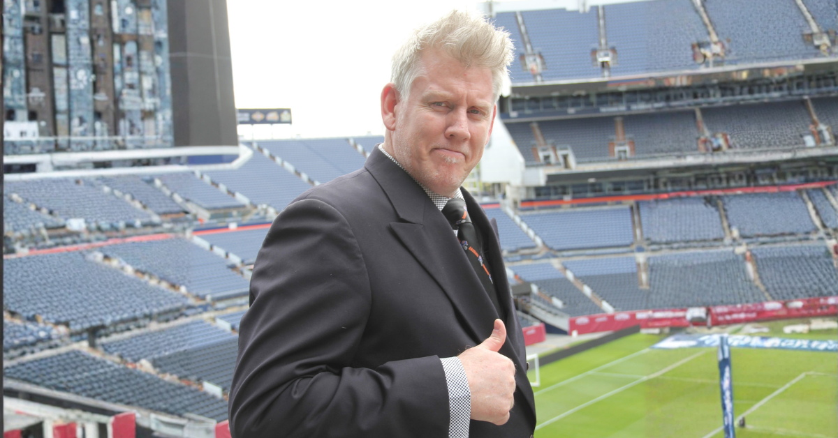 Man in a black suit giving a thumbs-up on a football stadium field with empty stands in the background.