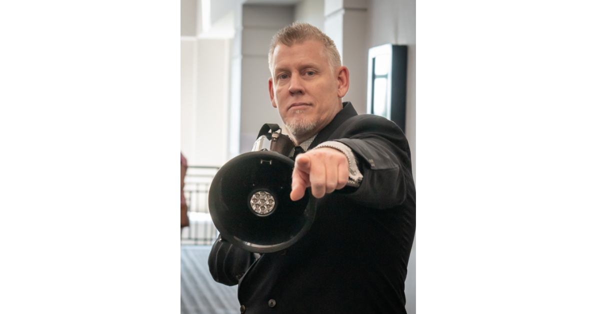 Man in a dark blazer points directly at the camera while holding a black circular device strapped to his arm.