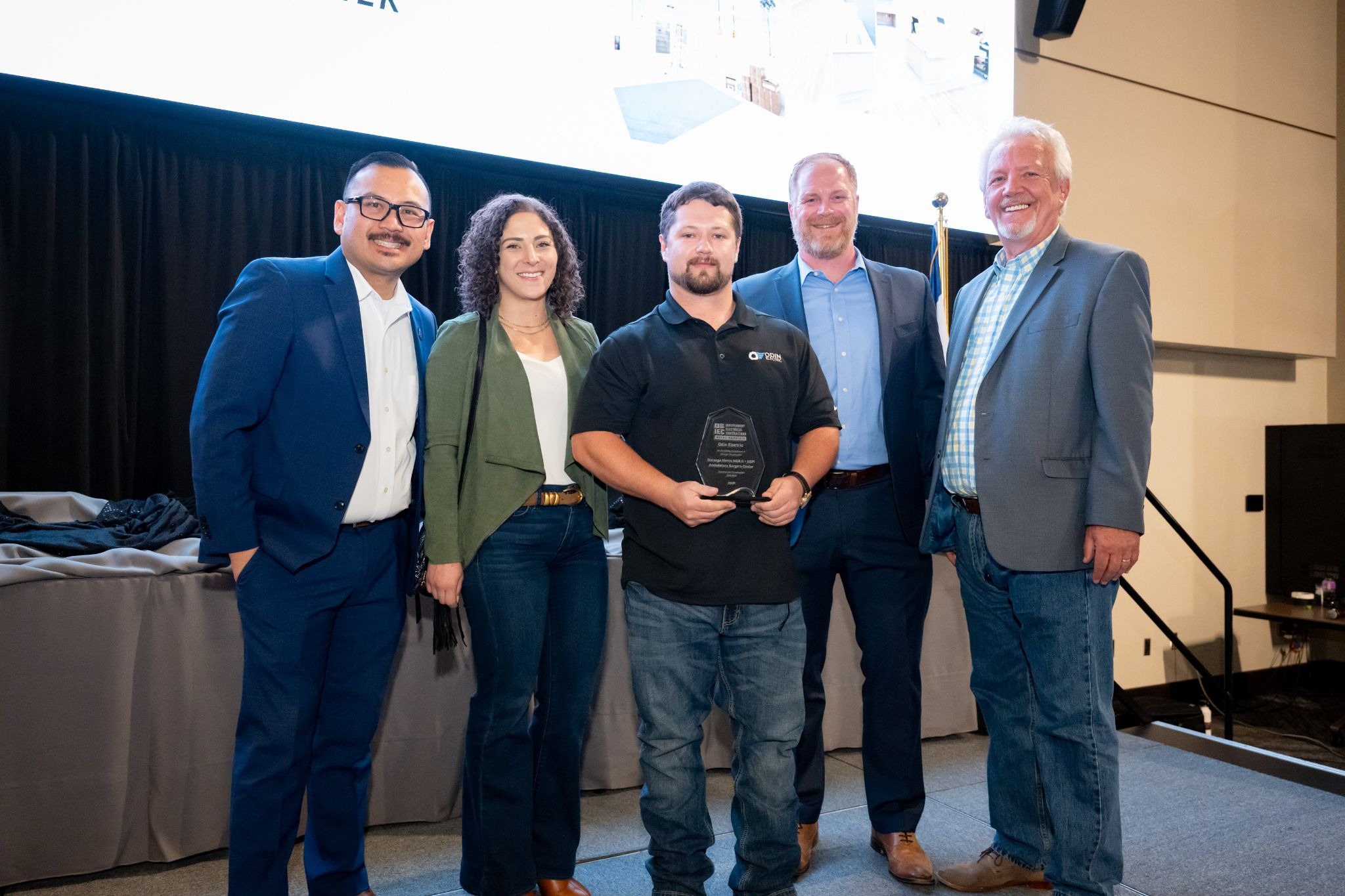 Five adults stand together on a stage, smiling for a photo after receiving an award; the man in the center holds a glass plaque.