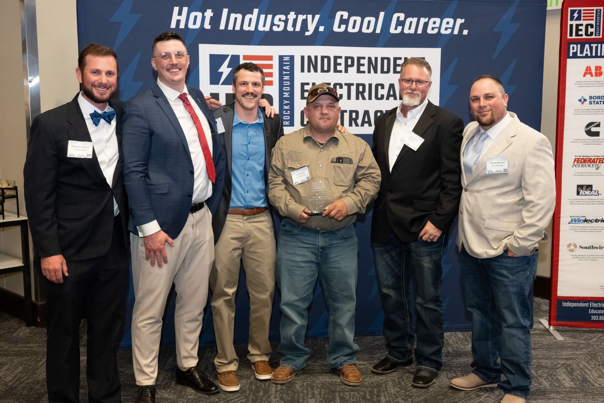 Six men in business and casual attire posing together at a trade show, with a blue backdrop and a banner reading 'Hot Industry. Cool Career.' behind them.