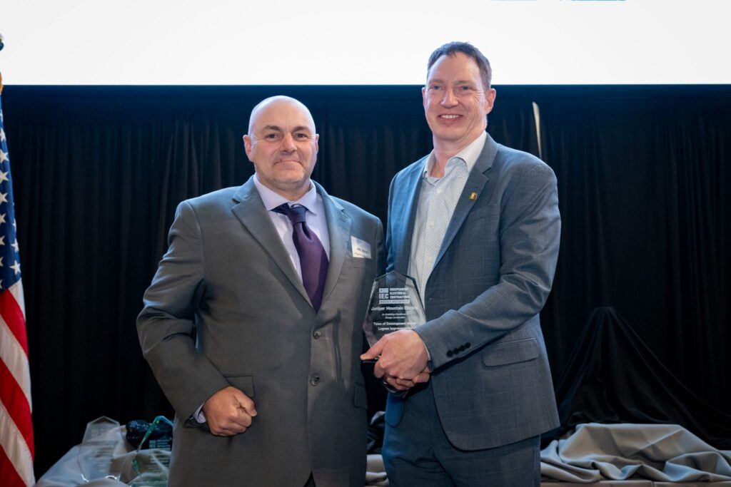 Two men in suits pose on a stage holding a glass award, with an American flag on the left and a black curtain backdrop behind them.
