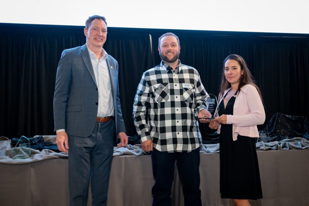 Three people on a stage pose for a photo; a man in a blue suit on the left, a man in a black-and-white plaid shirt center, and a woman in pink holding a trophy on the right.