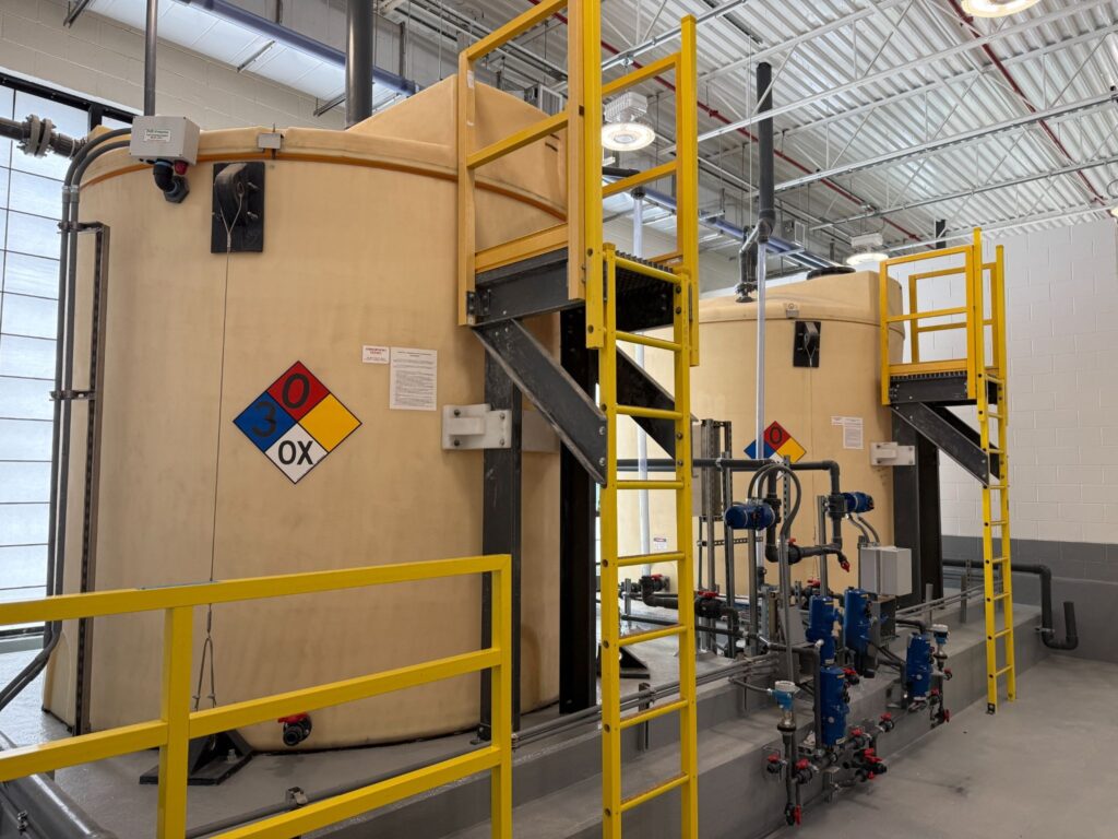 Industrial processing room with large beige storage tanks, yellow safety railings, and a network of pipes and valves.