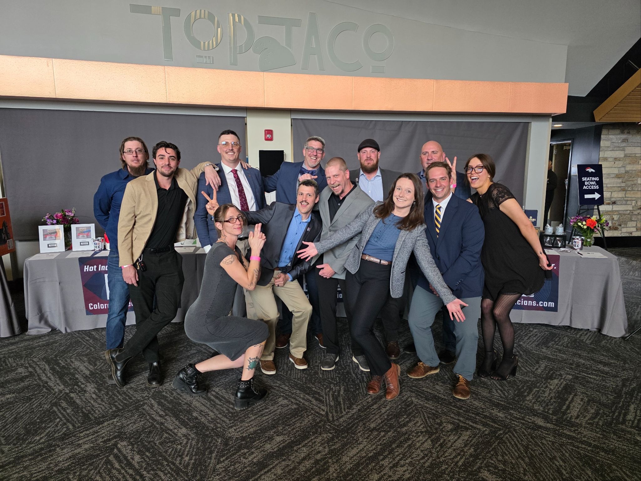 Group of adults in business attire posing together for a photo at an event, some making peace signs, in front of a backdrop and tables with flowers and displays.