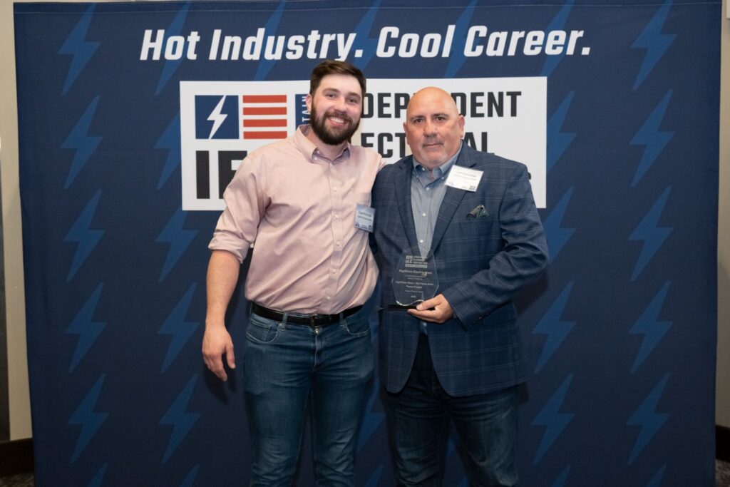 Two men pose together at an awards event; one holds a glass trophy while smiling at the camera, in front of a blue banner that says 'Hot Industry. Cool Career.'