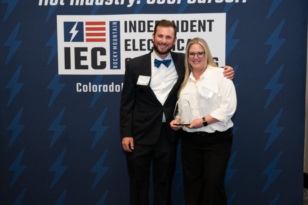 Man in a black suit and a woman in a white shirt pose together, holding glass awards at an IEC event in front of a blue backdrop with the IEC logo and 'Colorado' text (smiling, close together).