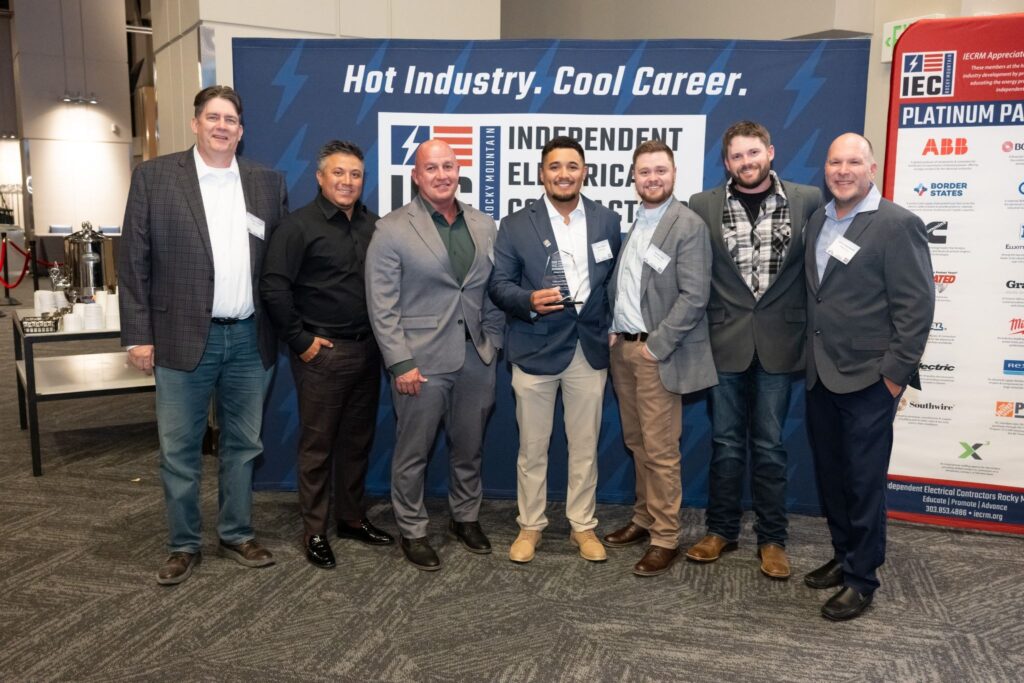 Seven men in suits pose for a photo at an IEC event, one holding a glass award in front of a blue banner with industry slogans.
