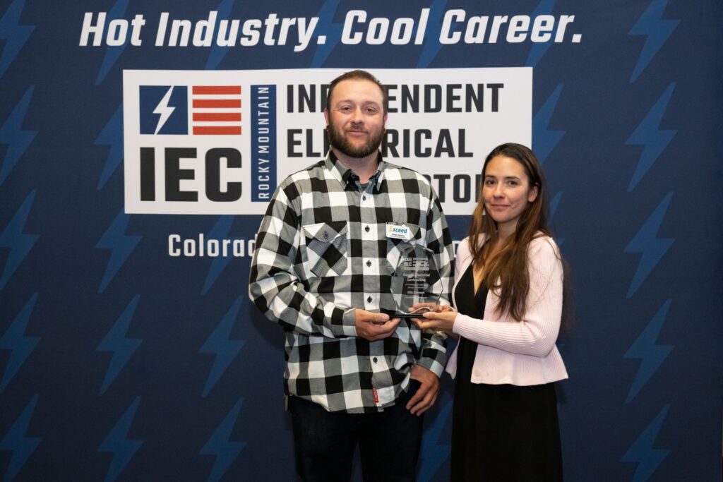 Two people stand side by side, a man in a black-and-white plaid shirt holding a glass award, with a woman in a light pink cardigan beside him, against a blue backdrop with logos and text.