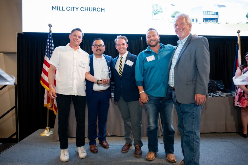 Five men in business casual posing on a stage; one holds a glass trophy, American flag behind them.