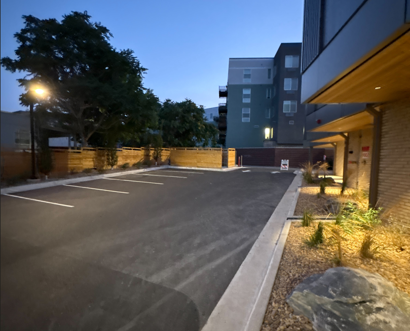 Empty parking lot at dusk beside a modern apartment building, with a wooden fence and street lamp nearby.