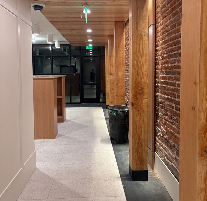Indoor corridor with brick wall on the right, wooden columns, and overhead lights leading to glass doors at the end; a trash can near the columns.