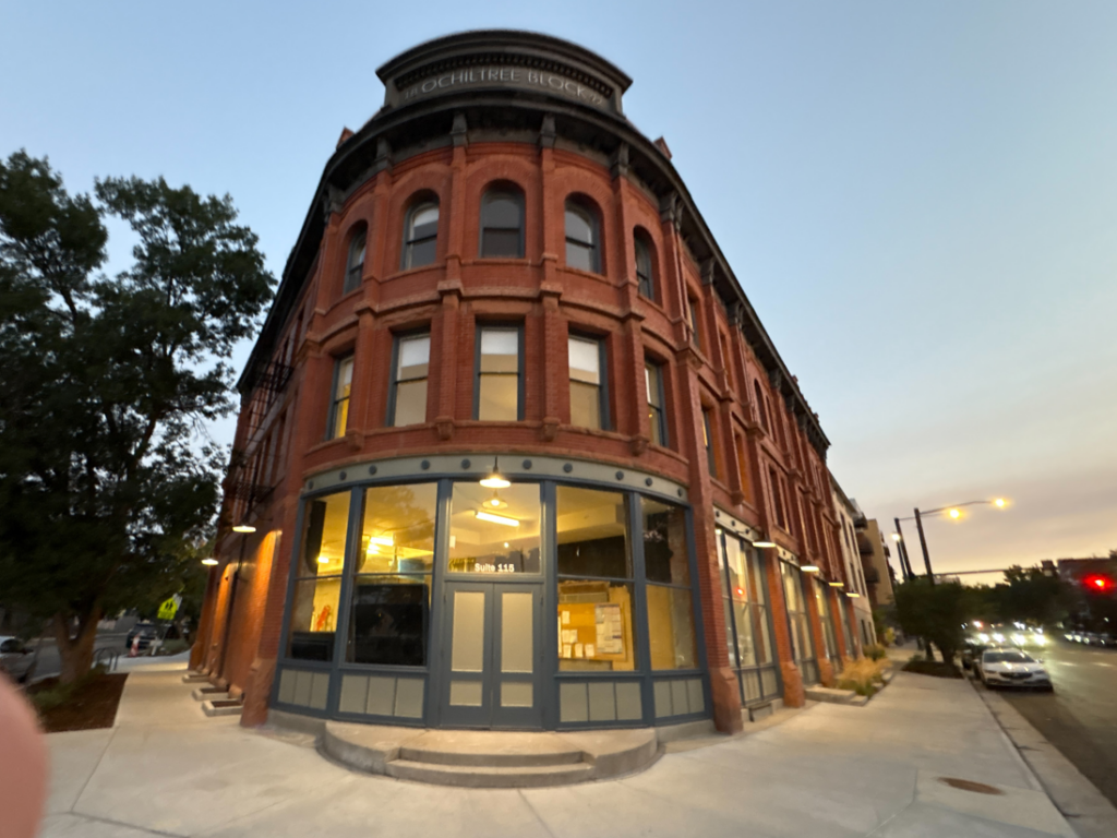 Red brick corner building with large storefront windows glowing from inside at dusk; street and trees to the left.
