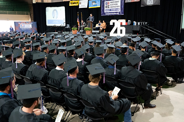 Graduation ceremony with hundreds of graduates in caps and gowns seated facing a stage where a speaker addresses them, banners overhead.