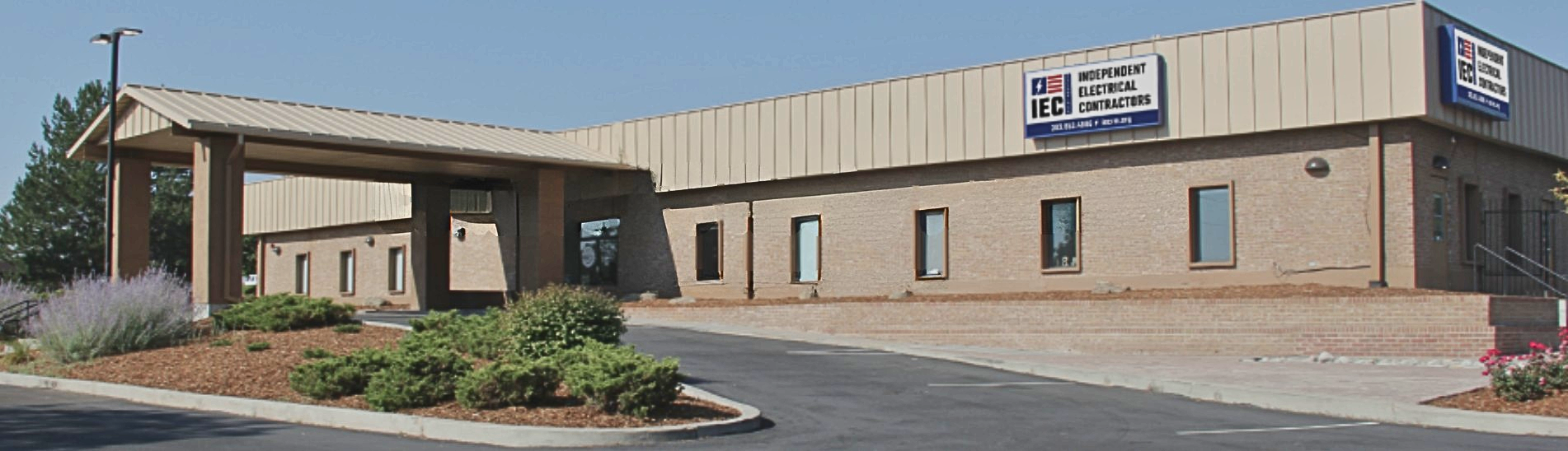Brick commercial building with a beige metal roof and a sign that reads Independent Electrical Contractors (IEC) at the front entrance area, with a curved driveway and landscaped front yard.